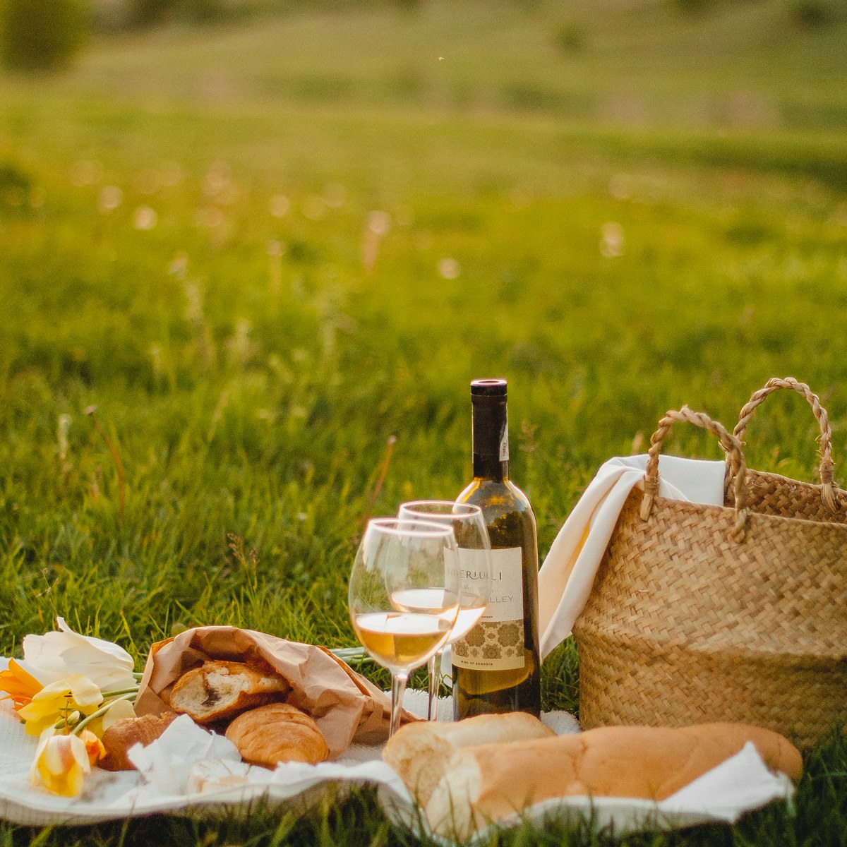 Open woven picnic tote beside bread and a bottle on grass